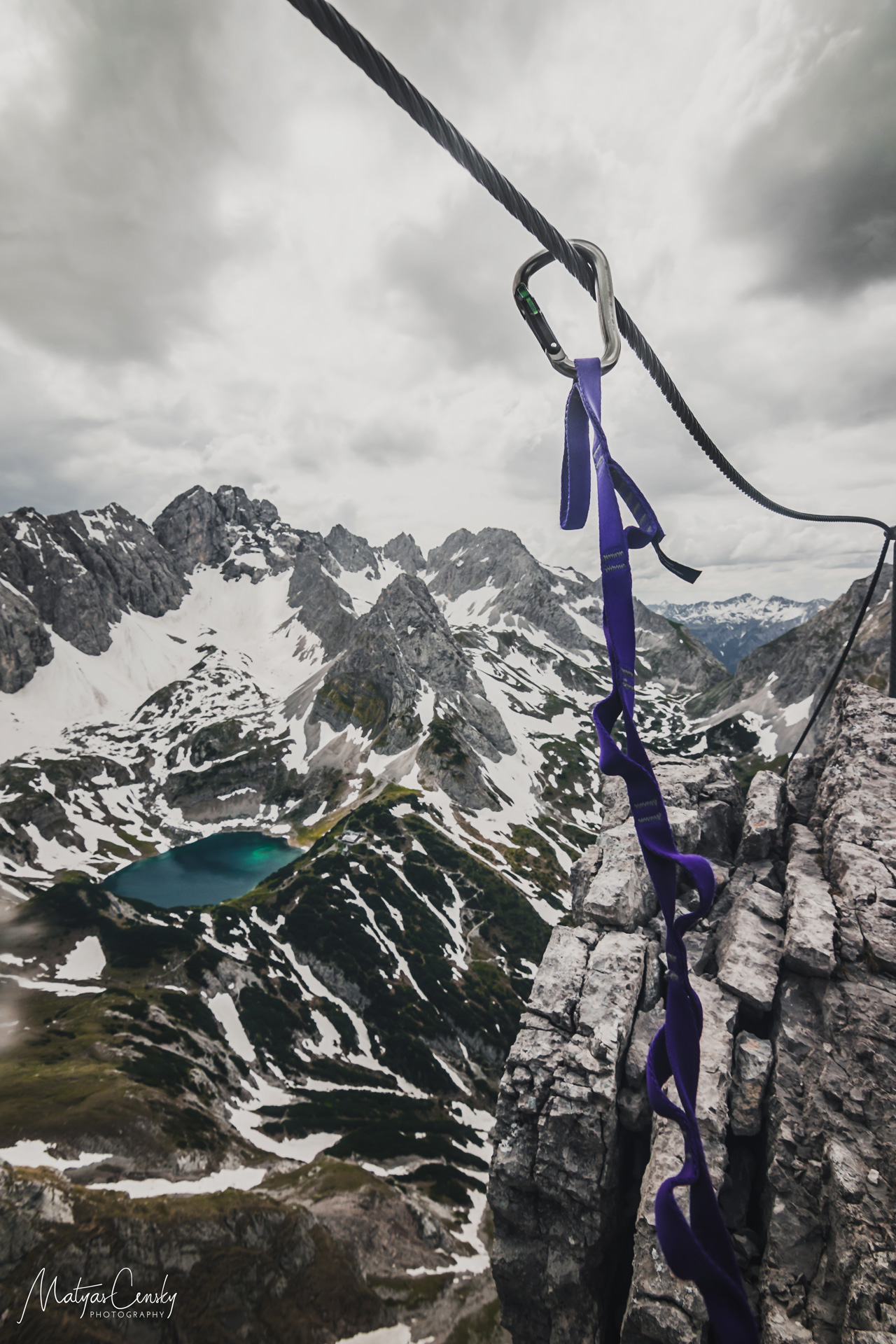Photo of a carabiner on a via ferrata rope with purple daisy chain as a protection with Drachensee, Austria in the background.