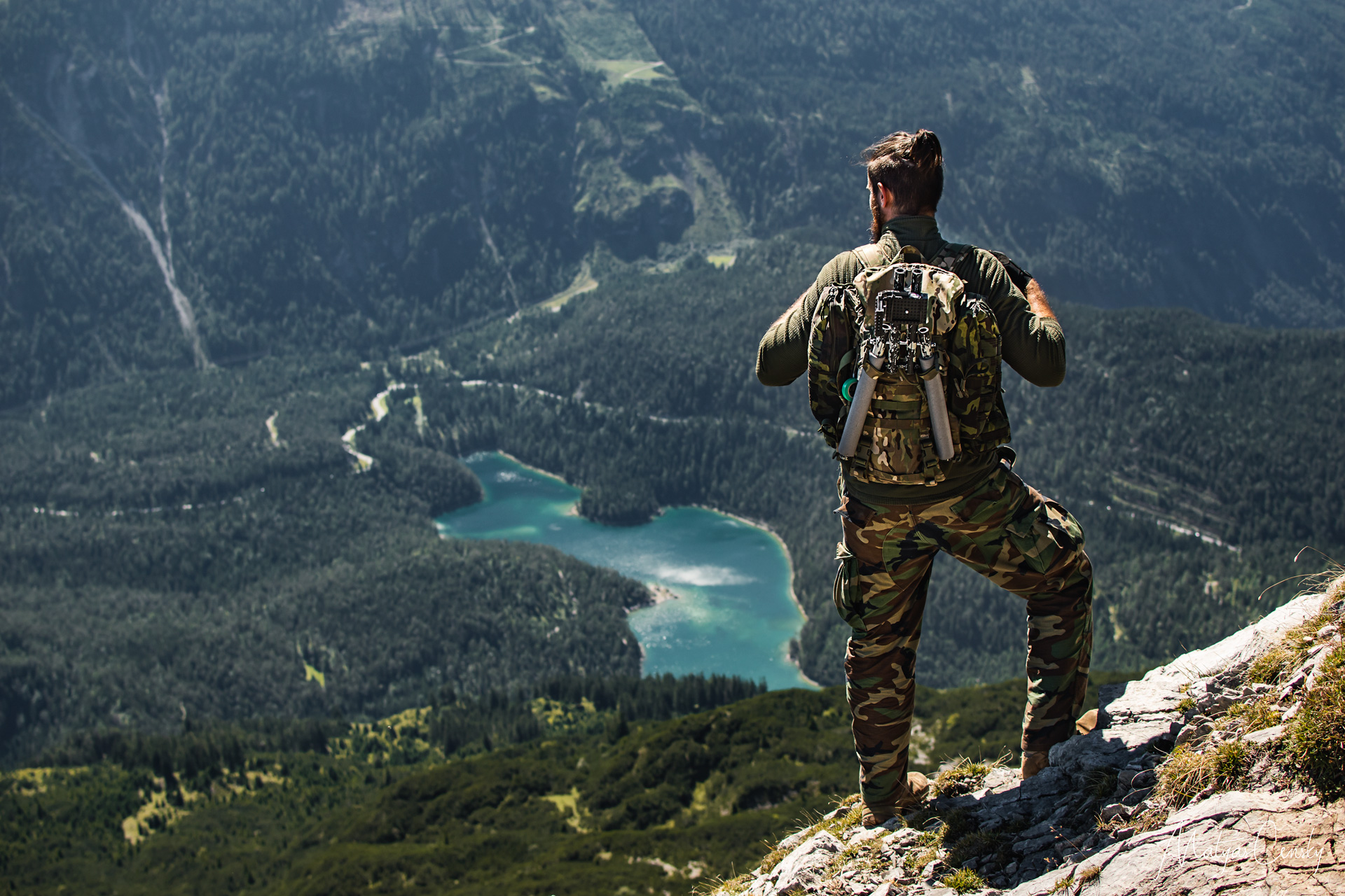 Photo of a men in camouflage outfit standing up in the mountains looking down on a blue lake.