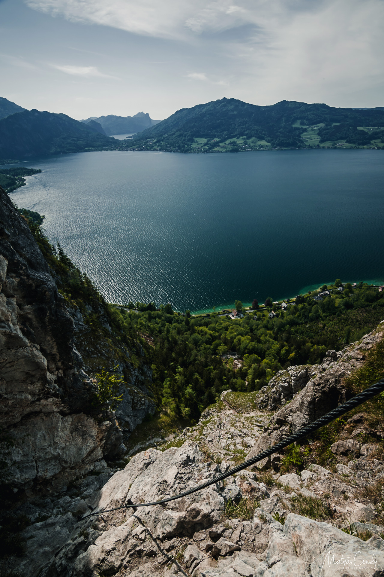 Landscape photo of Attersee in mid day from via ferrrata with a rock and fixed rope in the foreground and Mondsee in the background.