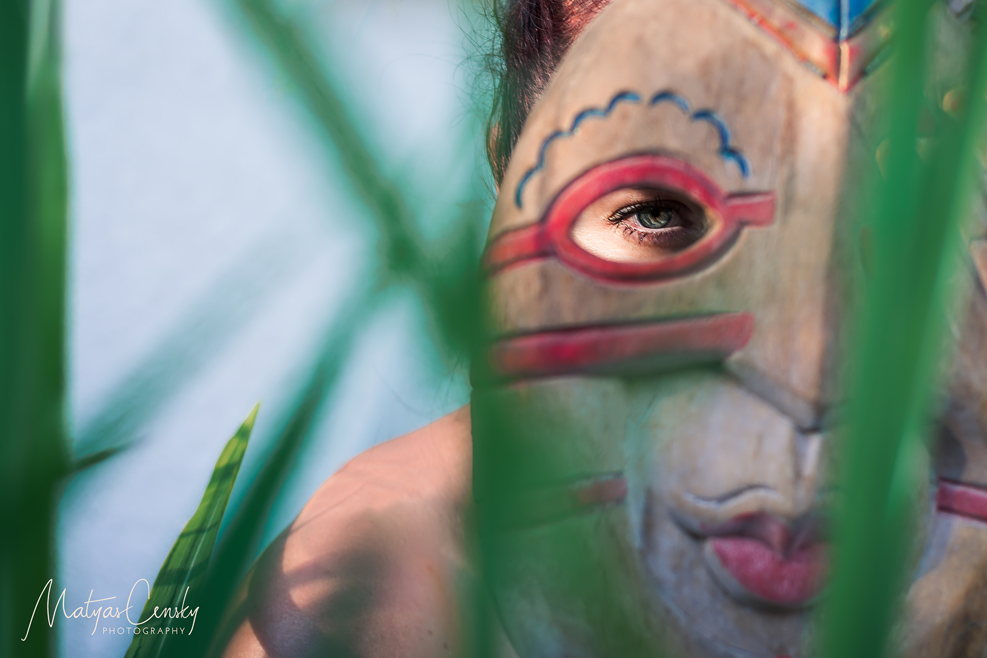 Photo of girl looking through wooden mask hiding behind green leaves focused on her eye through the mask.
