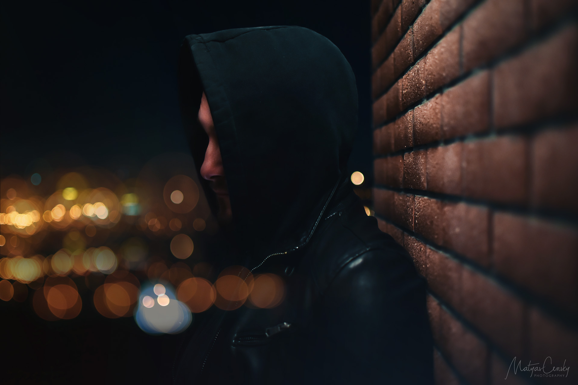 Portrait photo of Matyas Censky posing against a brick wall, in black leather jacket and hoody with night city lights blurred in back ground with bokeh effect.