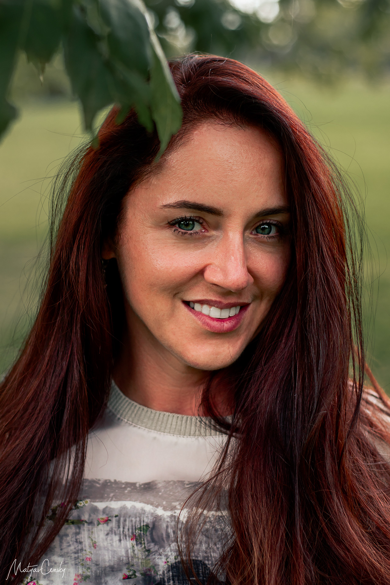 Headshot portrait photograph of a girl with dark red hair and green eyes smiling and standing under a tree in a park Stromovka in Prague.