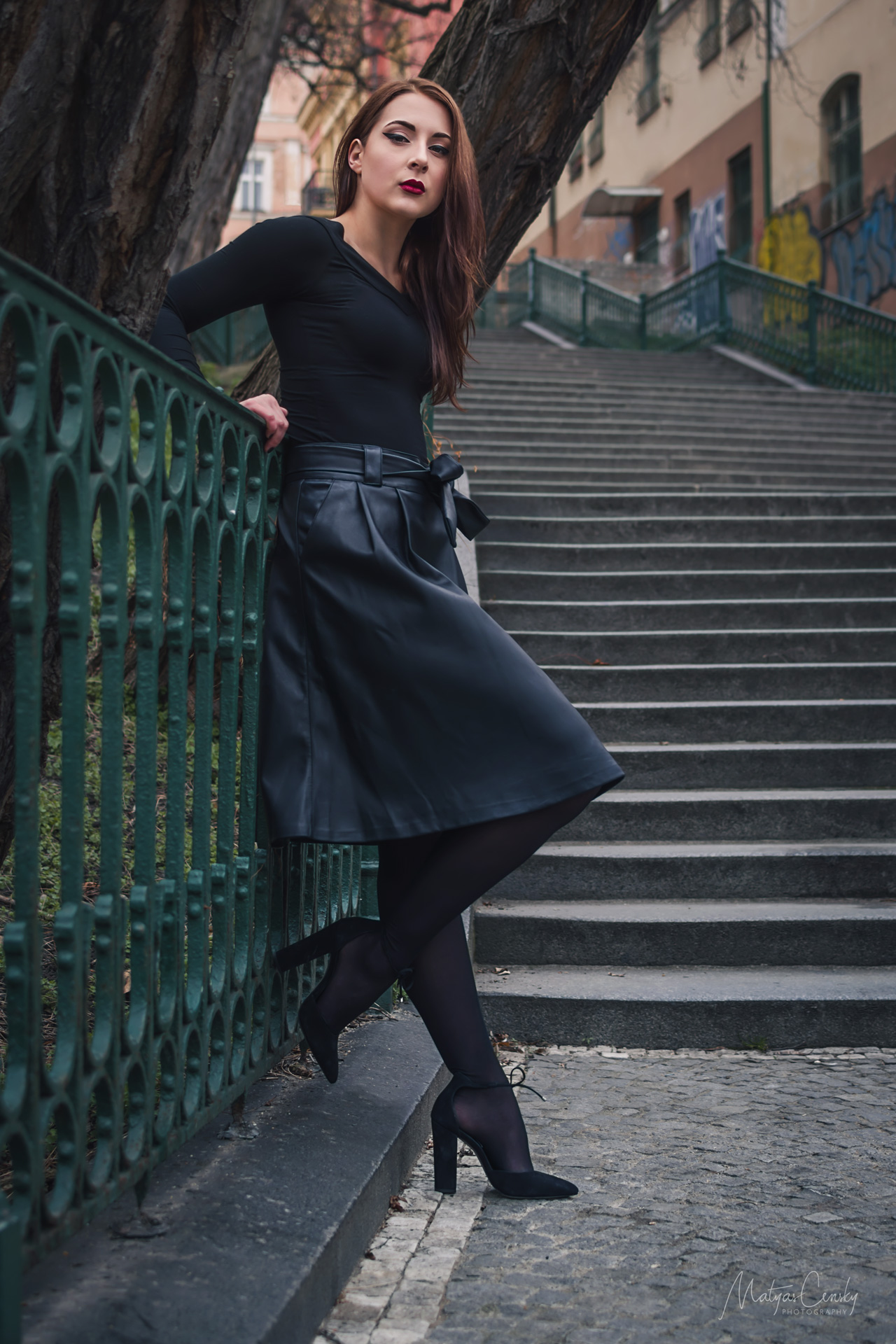 Full body shot of red hair model in black leather skirt, high heels, black top leaning against green decorative fence on stairs.