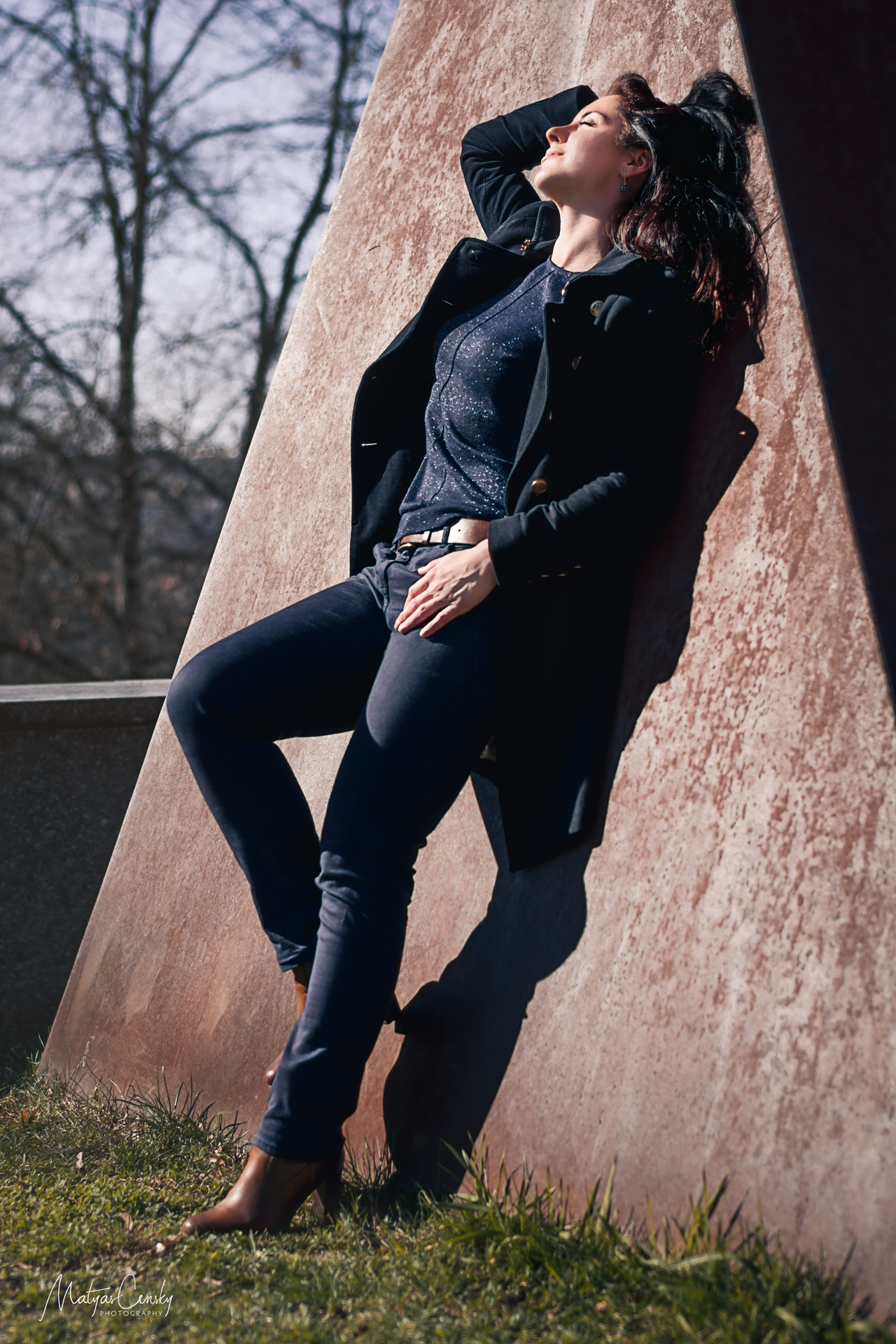Full body portrait photo of a girl in opened autumn coat, glitter top and blue jeans with brown heel shoes and brown belt leaning against iron sculpture facing and enjoying the sun.