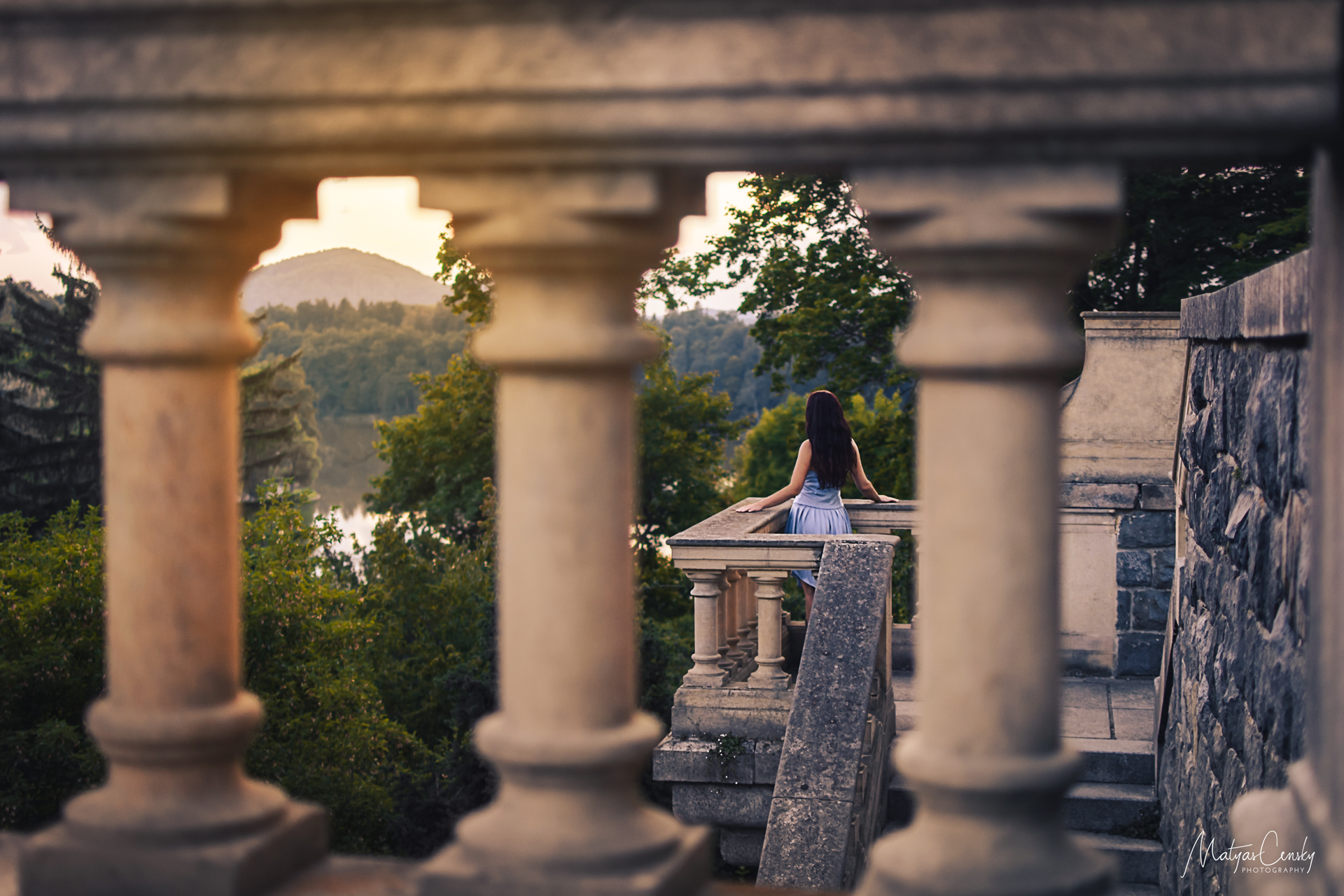 Photo of landscape shot through old stone railing with sunset behind a hill, lake and girl in blue dress watching the horizon at Konopiste park.