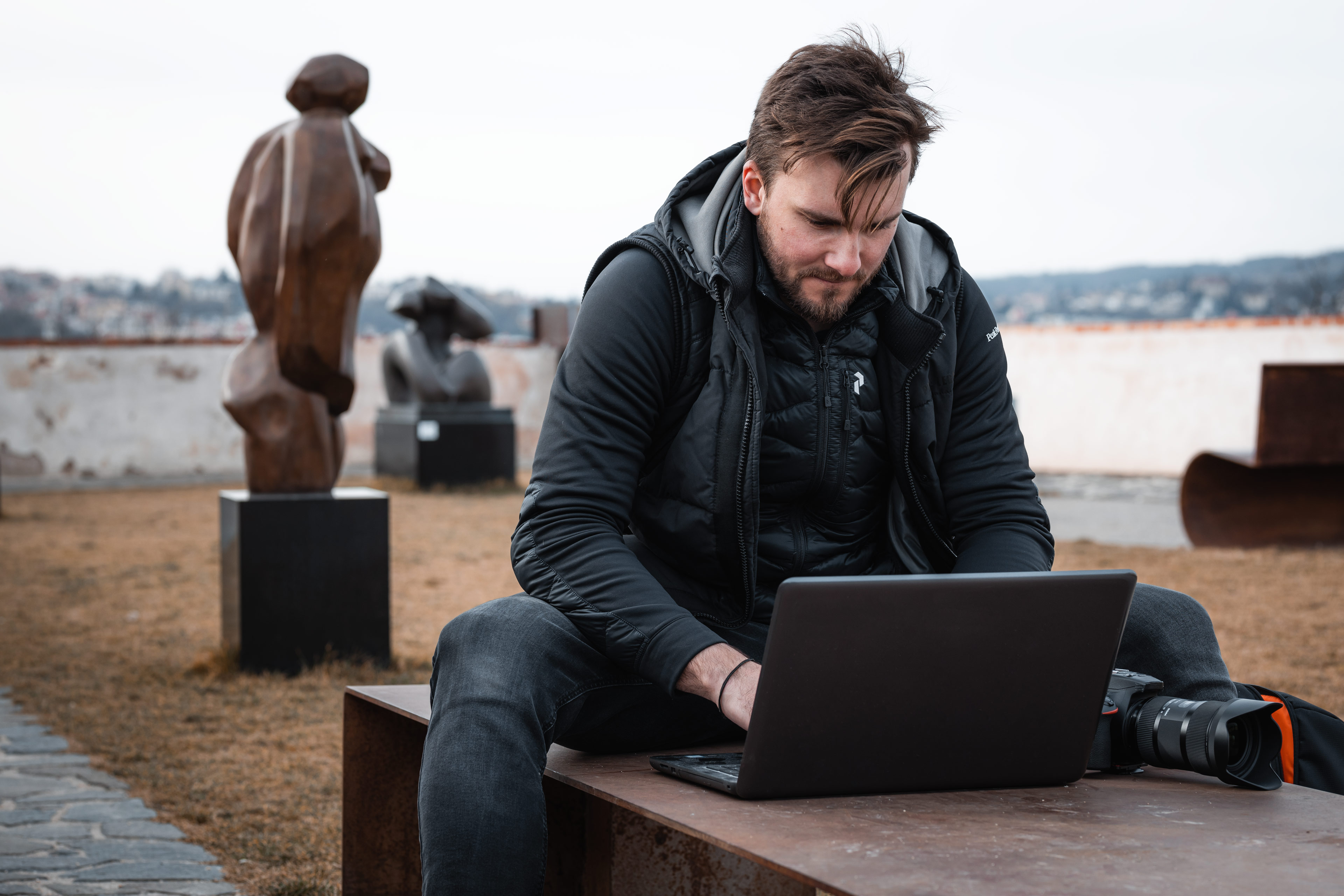 Matyáš Čenský sitting on a bench outside with a laptop and a camera working on his photography business.